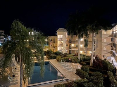 Nighttime view of a hotel courtyard with a rectangular pool, lounge chairs, and palm trees surrounding beige buildings.